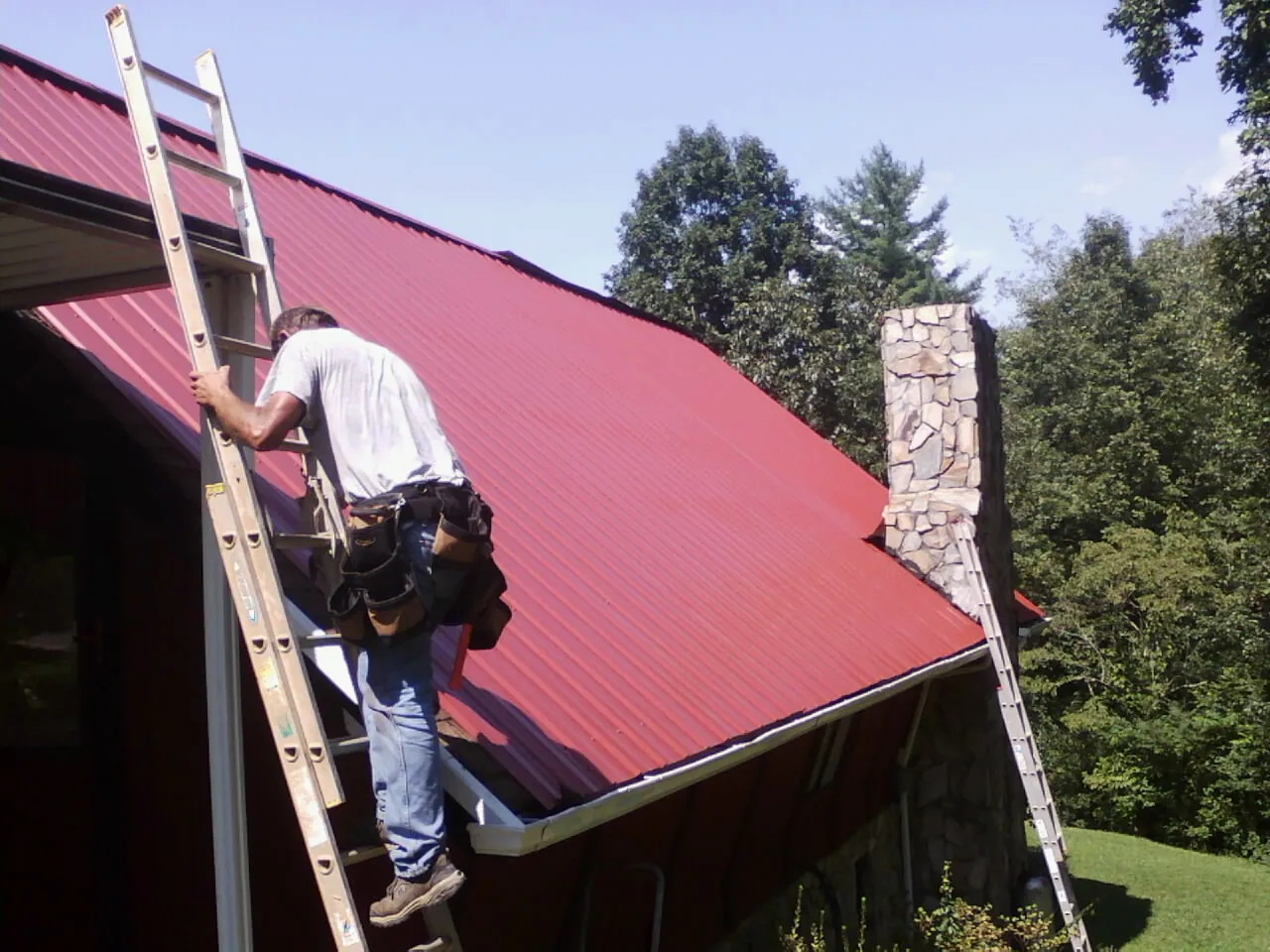 Roofer on a ladder beside a red metal roof and stone chimney — Soffit & Fascia Repair by Roofing Brothers in Keymar